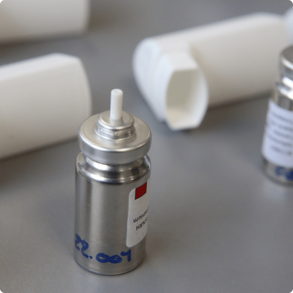 Two shiny metal pMDI inhaler cannisters on table with development codes beside white inhaler shell at Nanopharm.